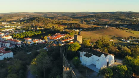 Drone-shot-of-a-white-chapel-building-on-a-hill-with-medieval-wall-and-tower-in-Alentejo,-Portugal