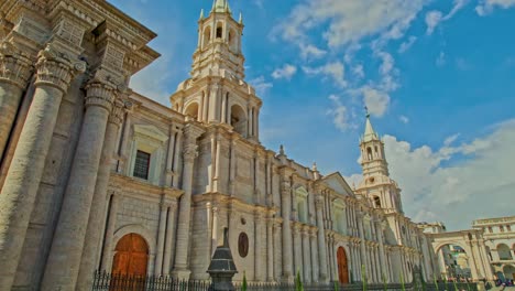 Drone-pan-of-Arequipa-Cathedral,-starting-from-the-left-portal-revealing-the-right-tower-and-the-entire-cathedral,-ending-with-a-local-tree,-on-a-sunny-day-with-scattered-clouds