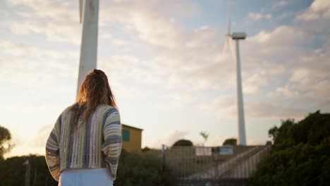 Slow-motion-shot-of-a-woman-walking-towards-windmills-turning-at-sunset