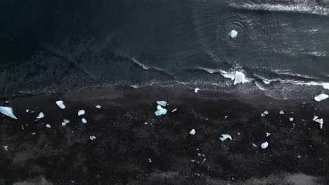 Aerial-view-from-Diamond-beach-in-the-summer-of-Iceland,-surrounded-by-black-sands-and-waves-with-incredible-icebers-over-the-beach-coast