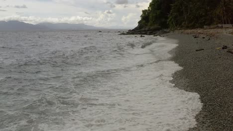 Coastal-Dolly-Shot-Along-the-Pebbled-Beach-with-Ocean-Waves-with-Scenic-Mountainous-Background-in-Looc-Bay,-Philippines