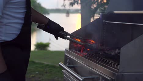 Close-up-of-griller-barbecue-grill-party-at-sunset-garden-lake-mexican-latin-service-cooker-flames-on-grid-and-turning-steak-meat-raw