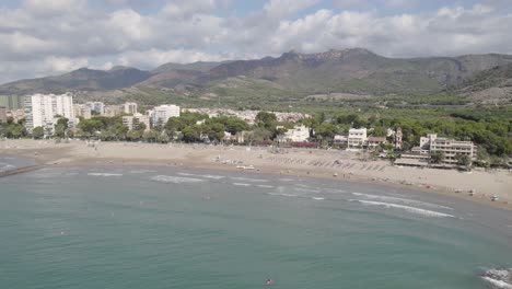 Lateral-aerial-view-of-the-Voramar-Hotel-and-beach-in-Benicassim-on-a-sunny-day-with-Desert-de-les-Palmes-mountains-in-the-background,-Spain