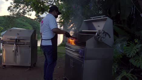 Latin-mexican-griller-taquero-grill-barbecue-with-flames-on-grid-preparing-raw-steak-meat-at-garden-lake-sunset-party-service