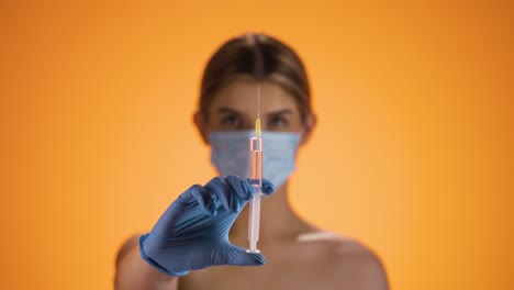 Medium-shot-of-a-young-pretty-nurse-wearing-a-ffp2-mask-while-holding-a-syringe-with-orange-liquid-into-the-camera-to-vaccinate-against-diseases-or-corona-virus-in-front-of-orange-background