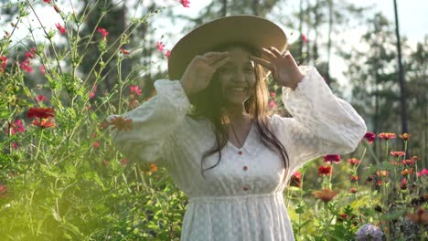 Little-girl-in-a-field-of-flowers,-wearing-hat-and-white-dress,-smiling-very-happy-during-sunset