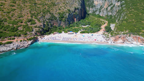 Aerial-drone-backward-moving-shot-over-turquoise-sea-water-of-the-Ionian-sea-with-sandy-beach-and-impressive-canyon-through-mountains-in-background