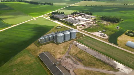 Aerial-view-of-agro-silos-tower-of-storage-of-agricultural-products-and-grain-elevators-in-the-middle-of-a-green-cultivated-field,-zoom-out