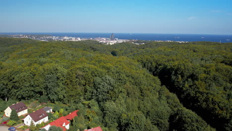Ascending-drone-shot-showing-suburb-houses,-forest-and-city-of-Gdynia-in-background---Blue-Baltic-Sea-at-horizon