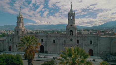 Aerial-drone-pan-from-left-to-right-showcasing-Arequipa-Cathedral-on-a-cloudy-day-with-intermittent-sunlight,-capturing-people-in-the-streets-and-local-vegetation-decorating-the-square
