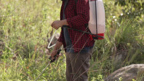 Medium-shot-of-a-man-working-with-a-backpack-sprayer-in-the-forest,-he-wears-a-red-shirt-and-a-hat