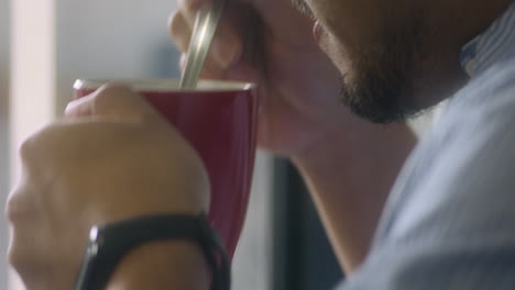 Close-Up-Shot-Of-A-Bearded-Man-Stirring-And-Drinking-His-Cup-Of-Coffee-In-A-Red-Mug
