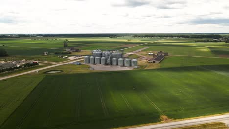 Aerial-view-of-agro-silos-tower-of-storage-of-agricultural-products-and-grain-elevators-in-the-middle-of-a-green-cultivated-field,-sliding-shot