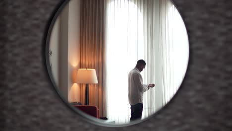 The-groom-standing-in-front-of-a-large-window-preparing-for-his-wedding,-with-his-back-reflected-in-a-round-mirror