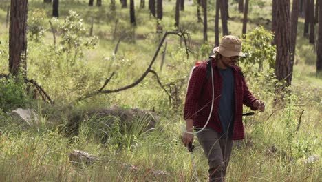 Medium-shot-of-a-forest-ranger-walking-with-a-backpack-sprayer-in-the-forest,-he-wears-a-red-shirt-and-a-hat