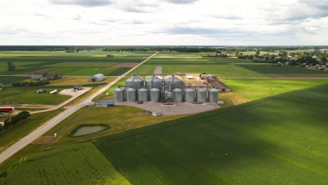 drone-view-of-agro-silos-tower-of-storage-of-agricultural-products-and-grain-elevators-in-the-middle-of-a-green-cultivated-field,-zooming-in