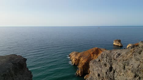 A-panoramic-aerial-view-captures-a-majestic-cliff,-a-rugged-rocky-beach,-and-a-distant-cargo-ship-on-the-horizon