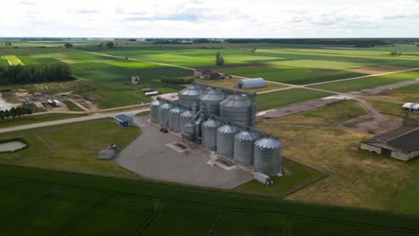 Aerial-view-of-agro-silos-granary-manufacturing-plant-storage-of-agricultural-products-and-grain-elevators-in-the-middle-of-a-green-cultivated-field,-parallax