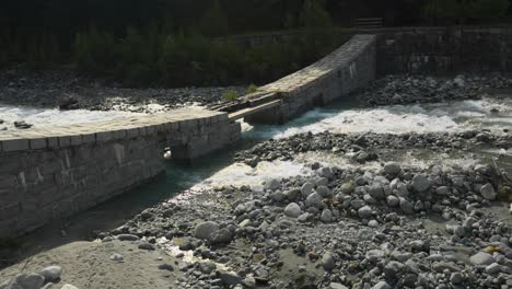 Stream-river-in-Italian-alps-passing-through-old-stone-bridge,-Alpe-ventina