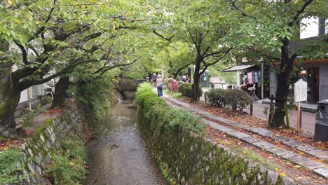 Vista-Panorámica-De-Los-Turistas-Caminando-Por-El-Camino-Del-Filósofo-Viajes-De-Kyoto-En-Japón,-Río-De-Verano-Con-Jardín-De-Follaje-Verde