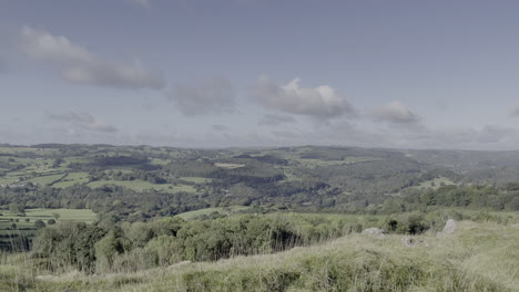 Eine-Reihe-Weißer-Windturbinen-Dreht-Sich-Auf-Einem-Hügel-Am-Horizont-Hinter-Sanften-Hügeln-Und-Tälern-In-Den-Derbyshire-Dales-An-Einem-Sonnigen-Tag-Mit-Blauem-Himmel-Und-Flauschigen-Wolken-Und-Breitem-Gras,-Das-Im-Wind-Weht