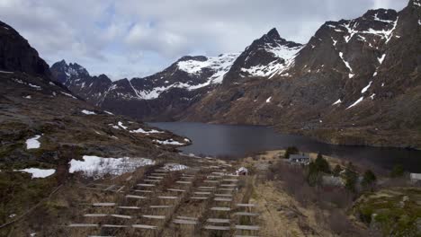 Ascending-drone-shot-of-a-view-towards-a-lake-and-dry-fish-rack-with-snow-capped-mountains-during-late-April,-A-in-Lofoten