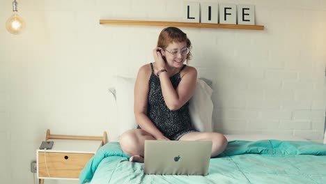 Young-woman-makes-a-video-call-on-laptop,-interacts-smiles-sitting-on-bed-in-bright-bedroom
