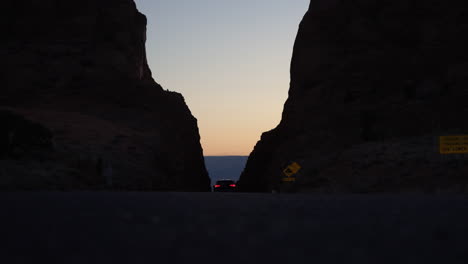 Car-driving-down-desert-road-through-two-big-mountainsides-at-sunset