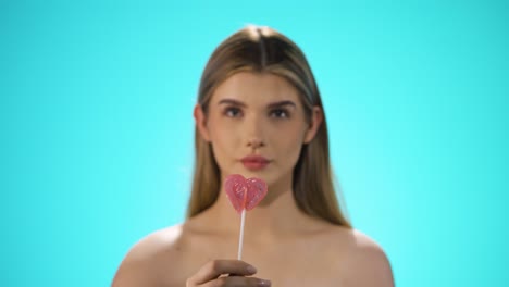 Static-medium-shot-of-a-young-pretty-blonde-woman-holding-a-lollipop-to-the-camera-and-then-taking-it-with-relish-while-she-licks-it-and-has-a-smile-on-her-lips-in-front-of-turquoise-background