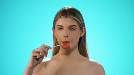 Static-shot-of-a-young-pretty-blonde-model-while-she-licks-a-sweet-lollipop-in-heart-shape-with-pleasure-and-looks-neutrally-to-the-camera-in-front-of-turquoise-background