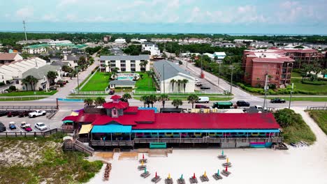 Pompano-Joe's-restaurant-and-parking-lot-trucking-right-aerial-drone-shot-with-a-view-of-old-98,-white-sand,-emerald-green-water-and-lots-of-umbrellas-and-beach-chairs-in-Destin-Florida
