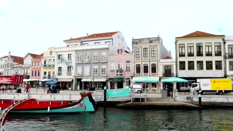 Panning-left-shot-capturing-Moliceiro-boats-in-front-of-traditional-buildings-along-a-canal-in-Aveiro,-Portugal