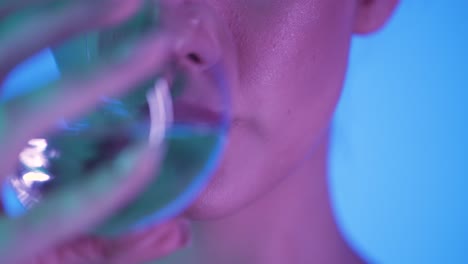 Close-up-shot-of-a-young-woman-with-full-beautiful-lips-drinking-a-glass-of-water-to-quench-her-thirst-against-blue-background-with-purple-contrast-in-her-face-in-slow-motion