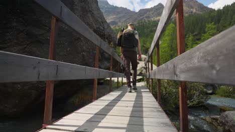 Vista-Trasera-De-ángulo-Bajo-Del-Excursionista-Masculino-Cruzando-Un-Puente-De-Madera-En-La-Cordillera