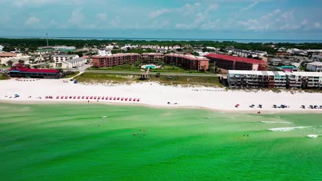 Destin-FL-drone-shot-flying-sideways-over-the-Gulf-of-Mexico-with-a-view-of-the-white-sand,-emerald-green-water,-pompano-joes-restaurant,-golf-cart,-parking-lot,-beach-chairs,-and-umbrellas-2-of-2