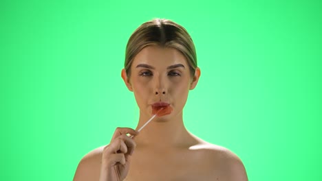 Static-slow-motion-shot-of-a-blonde-young-woman-licking-a-heart-shaped-lollipop-and-letting-the-sugar-taste-melt-on-her-lips-in-front-of-green-background