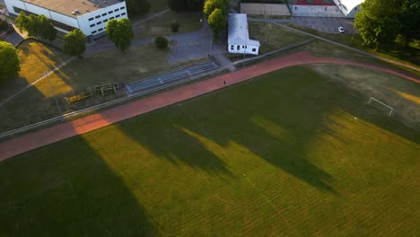 drone-shot-of-a-green-football-field-during-the-sunset,-there-is-runner-who-runs-around-the-stadium