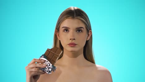 Medium-shot-of-a-young-attractive-woman-while-she-enjoys-a-bite-of-the-vegan-chocolate-bar-and-afterwards-strokes-her-lips-with-her-finger-in-front-of-turquoise-background-in-slow-motion