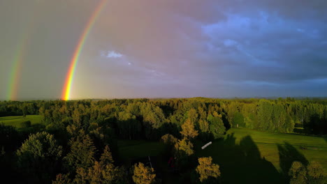 Aerial-drone-tilt-up-shot-of-two-rainbows-falling-into-mystic-forest-along-with-rural-cottages-after-rainfall