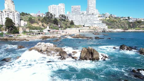 Ocean-Waves-Hitting-the-Coastal-Rocks-in-Viña-del-Mar-with-Buildings-in-the-Background,-Chile