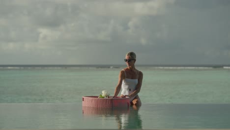 Woman-sitting-on-infinity-pool-edge-enjoying-drink-from-floating-breakfast