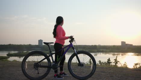 Woman-stands-with-her-bike-while-she-takes-off-her-helmet-and-looks-out-over-a-lake