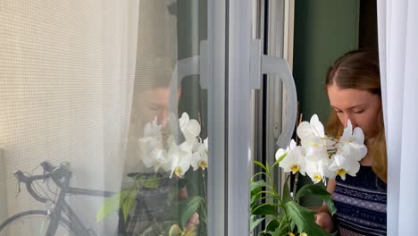 Junge-Frau-Gießt-Weiße-Blumen-Am-Fenster-Zu-Hause