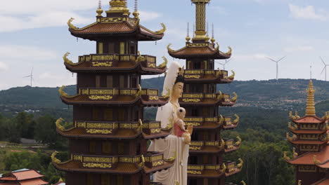 Luftaufnahme-Eines-Buddhistischen-Tempels-Mit-Statue-In-Vietnam-Und-Rotierenden-Windkraftanlagen-Auf-Einem-Hügel-Im-Hintergrund