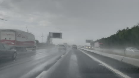 Vehicles-kick-up-water-spray-as-they-pass-white-wind-turbines-while-traveling-along-a-motorway-during-a-heavy-rain-storm