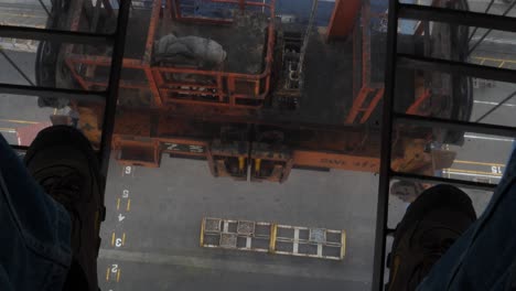 Crane-operator-POV-looking-through-glass-floor-at-the-ground-and-the-shipment-cargo-from-height-dangerously-high