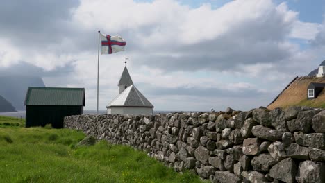 Wide-shot-showing-waving-faroese-flag-in-front-of-small-church-on-Faroe-Islands-during-cloudy-day