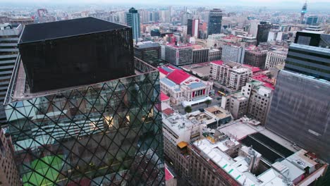 Absteigende-Luftaufnahme-Eines-Glasgebäudes-In-Einem-Rautenmuster-In-Der-Innenstadt-Von-Santiago,-Chile,-Mit-Dem-Stadttheater-Im-Hintergrund