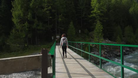 Female-hiker-crosses-sunny-mountain-river-on-wooden-bridge