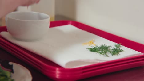 Ein-Zweig-Dill-Und-Zitronenschale-Auf-Einem-Weißen-Papiertuch,-Rote-Mittagstabletts-Und-Eine-Weiße-Glasschüssel,-Während-Ein-Koch-Im-Vordergrund-Eine-Mahlzeit-Zubereitet